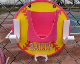 Peut inclure: Un support de softball personnalisé avec un design jaune et rose. Le support porte le nom "BAILEY" en lettres blanches. Il comporte un centre rose et des crochets blancs pour l'accrochage. Le support est fixé à une clôture en treillis métallique.
