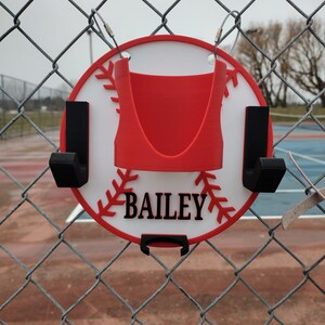 May include: A baseball-themed sign with the name "BAILEY" in black lettering. The sign is red, white, and black, and is designed to hang on a chain-link fence. The sign has a red baseball stitch design around the edge.