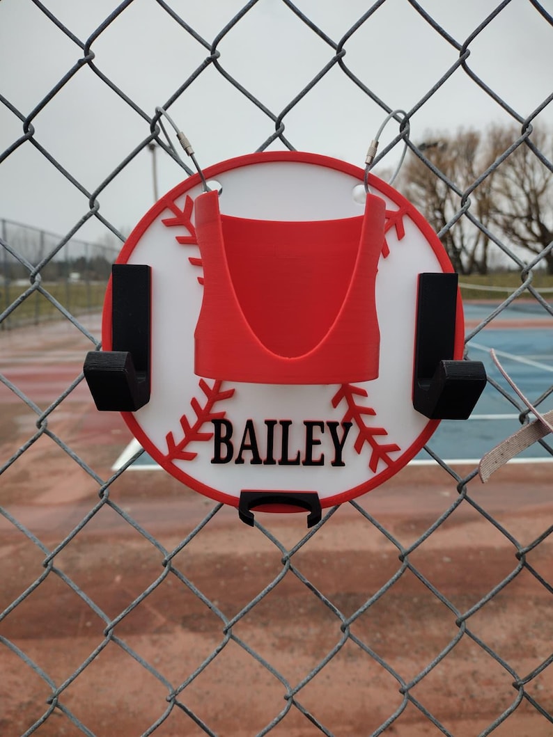 May include: A baseball-themed sign with the name "BAILEY" in black lettering. The sign is red, white, and black, and is designed to hang on a chain-link fence. The sign has a red baseball stitch design around the edge.