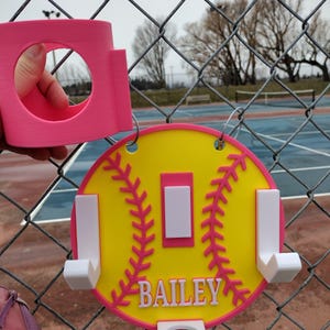 May include: A pink and yellow softball-themed sign with the name "BAILEY" in white lettering. The sign has a pink border and stitching detail, with white hooks and a pink cup holder. The sign is hanging on a chain-link fence.