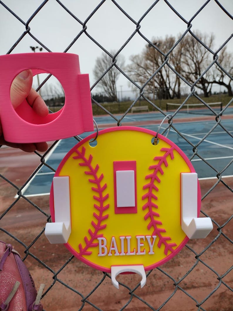 May include: A pink and yellow softball-themed sign with the name "BAILEY" in white lettering. The sign has a pink border and stitching detail, with white hooks and a pink cup holder. The sign is hanging on a chain-link fence.