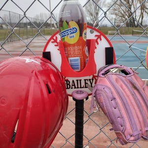 May include: A red baseball helmet, a pink and black baseball bat, and a pink baseball glove hang on a chain-link fence. A red holder with a bottle of Snapple peach tea is attached to a baseball-themed sign that reads "BAILEY".