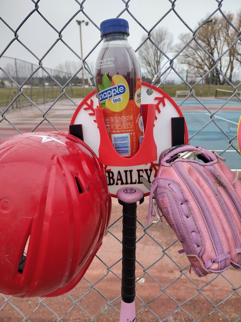 May include: A red baseball helmet, a pink and black baseball bat, and a pink baseball glove hang on a chain-link fence. A red holder with a bottle of Snapple peach tea is attached to a baseball-themed sign that reads "BAILEY".