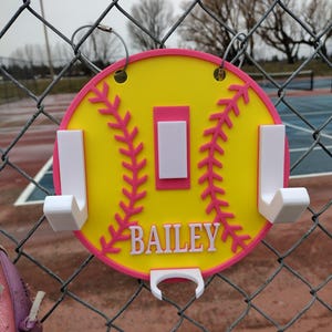 May include: A yellow and pink softball-shaped wall hook with the name "BAILEY" in white. The hook has a pink border and stitching details, with white hooks for hanging items. It is hanging on a chain-link fence.