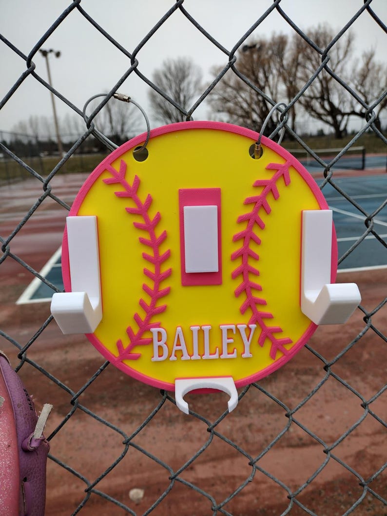 May include: A yellow and pink softball-shaped wall hook with the name "BAILEY" in white. The hook has a pink border and stitching details, with white hooks for hanging items. It is hanging on a chain-link fence.
