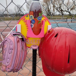 May include: A baseball-themed setup featuring a pink bat, a red helmet, and a pink and purple glove. A bottle of Snapple tea is held in a pink holder attached to a yellow baseball-shaped sign with the name "BAILEY". The items are displayed on a chain-link fence.