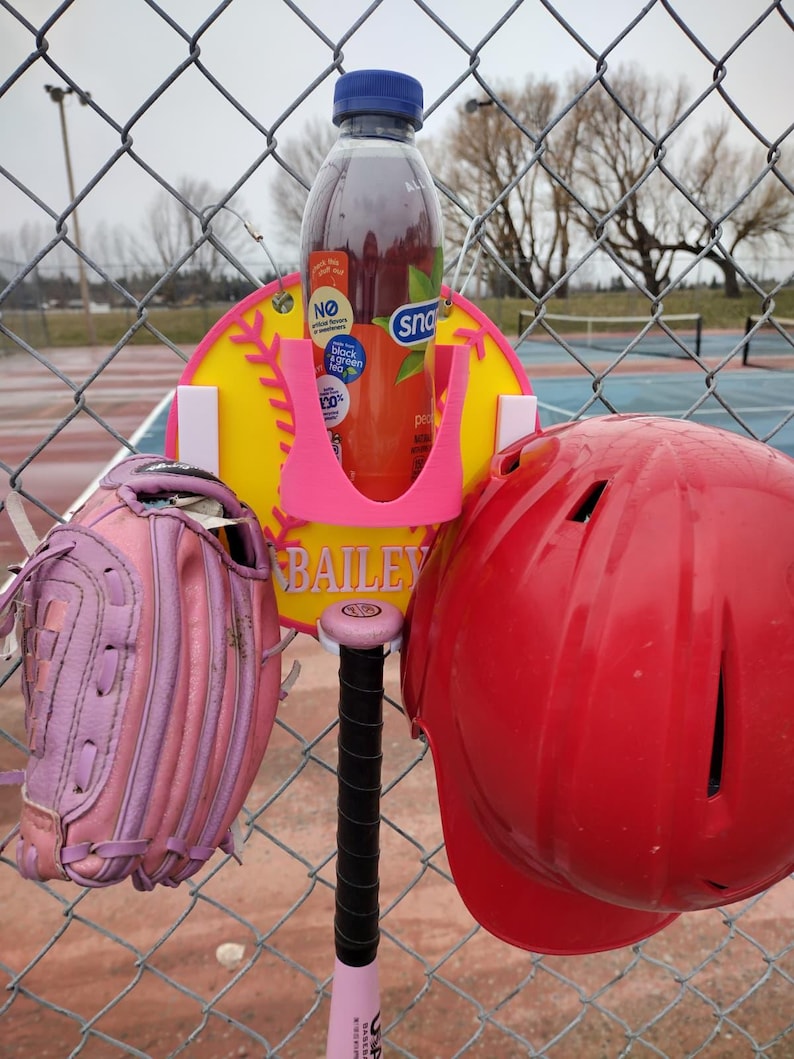 May include: A baseball-themed setup featuring a pink bat, a red helmet, and a pink and purple glove. A bottle of Snapple tea is held in a pink holder attached to a yellow baseball-shaped sign with the name "BAILEY". The items are displayed on a chain-link fence.