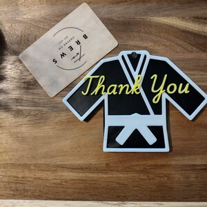 May include: A black and white karate uniform-shaped ornament with the words "Thank You" in yellow script. A small, rectangular card with the text "Brews Coffee Co." is also present.