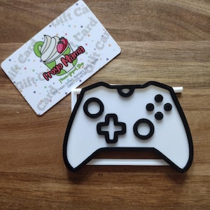 May include: A white and black plastic video game controller shaped cookie cutter. Next to it is a white gift card with a logo featuring a green frozen yogurt swirl, a red strawberry, and the text Froyo Mama, Frozen Yogurt Shop, 7206 Main Street.