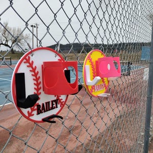 May include: Two baseball-themed sports organizers are attached to a chain-link fence. One is white with red accents and the name "BAILEY". The other is yellow with pink accents and the name "BAILEY". Both have a cup holder and hooks.