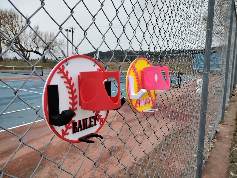 May include: Two baseball-themed sports organizers are attached to a chain-link fence. One is white with red accents and the name "BAILEY". The other is yellow with pink accents and the name "BAILEY". Both have a cup holder and hooks.