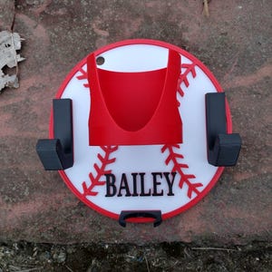 May include: A baseball-themed wall mount with a red and white design. The mount features a red holder, two black hooks, and the name "Bailey" in black lettering. The design resembles a baseball with red stitching.