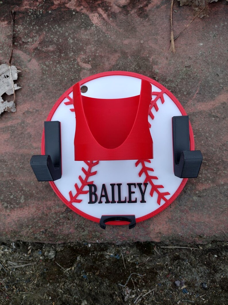 May include: A baseball-themed wall mount with a red and white design. The mount features a red holder, two black hooks, and the name "Bailey" in black lettering. The design resembles a baseball with red stitching.