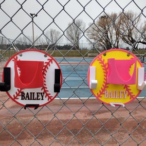 May include: Two baseball-themed wall organizers, one red and white, the other yellow and pink, are displayed on a chain-link fence. Each has a holder and hooks, with the name "BAILEY" in white.