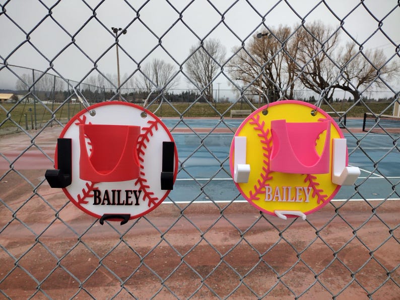 May include: Two baseball-themed wall organizers, one red and white, the other yellow and pink, are displayed on a chain-link fence. Each has a holder and hooks, with the name "BAILEY" in white.