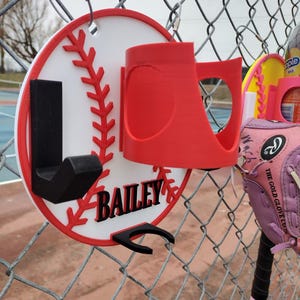May include: A baseball-themed holder with the name "BAILEY" in black lettering. The holder is red, white, and black, designed to hang on a chain-link fence. A red cup holder and black hook are attached to the baseball design.