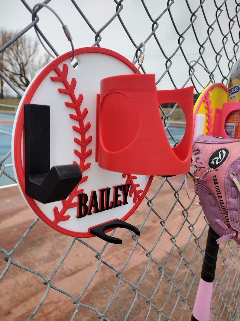 May include: A baseball-themed holder with the name "BAILEY" in black lettering. The holder is red, white, and black, designed to hang on a chain-link fence. A red cup holder and black hook are attached to the baseball design.