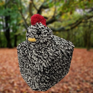 May include: A black and white fluffy chicken toy with a red comb and yellow beak. The toy is sitting on a bed of brown leaves.