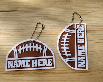 May include: Two brown and white silicone keychains shaped like footballs, each featuring a white lace design and a rectangular space with the text NAME HERE. Both items include a silver-colored metal ball chain for attachment to keys or bags.