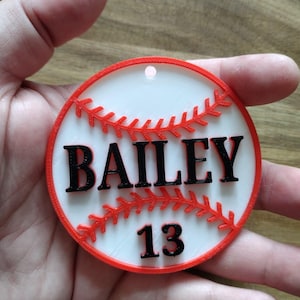 以下が含まれることがあります： 白い中心と赤い縁取りの野球をテーマにしたオーナメント。「BAILEY」の文字と数字の「13」が黒で印刷されています。オーナメントの上部には、吊り下げ用の小さな穴があります。