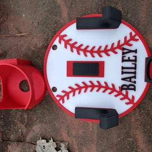May include: A red baseball-themed wall mount and a separate red holder. The wall mount is white with red stitching and a red border, featuring the name "BAILEY" in black. The holder is a rounded shape with a circular opening.