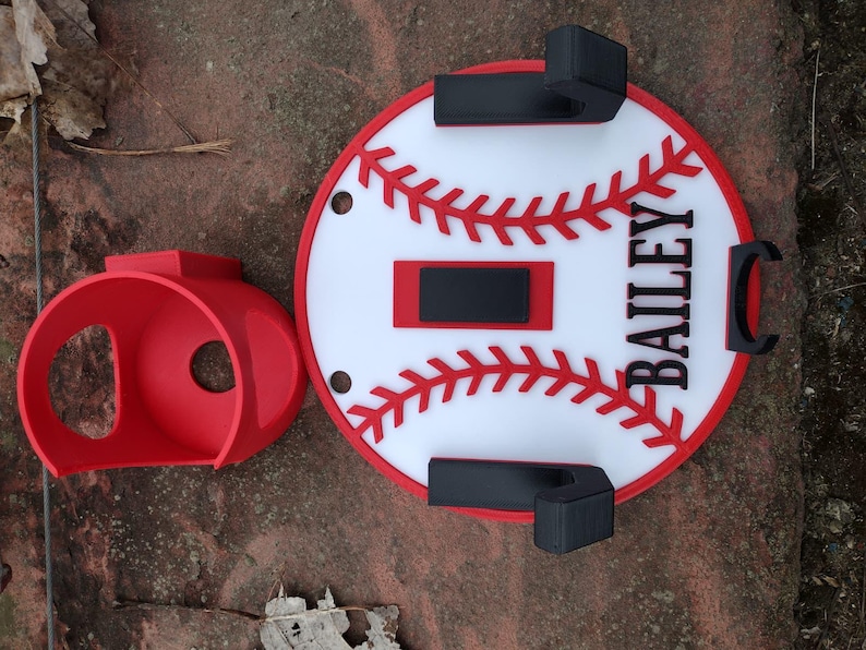 May include: A red baseball-themed wall mount and a separate red holder. The wall mount is white with red stitching and a red border, featuring the name "BAILEY" in black. The holder is a rounded shape with a circular opening.