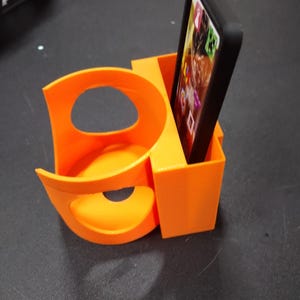 May include: Bright orange 3D-printed car cup holder and phone holder. The cup holder has a curved design with a circular opening. The phone holder is a rectangular box, holding a black phone with a screen displaying app icons.