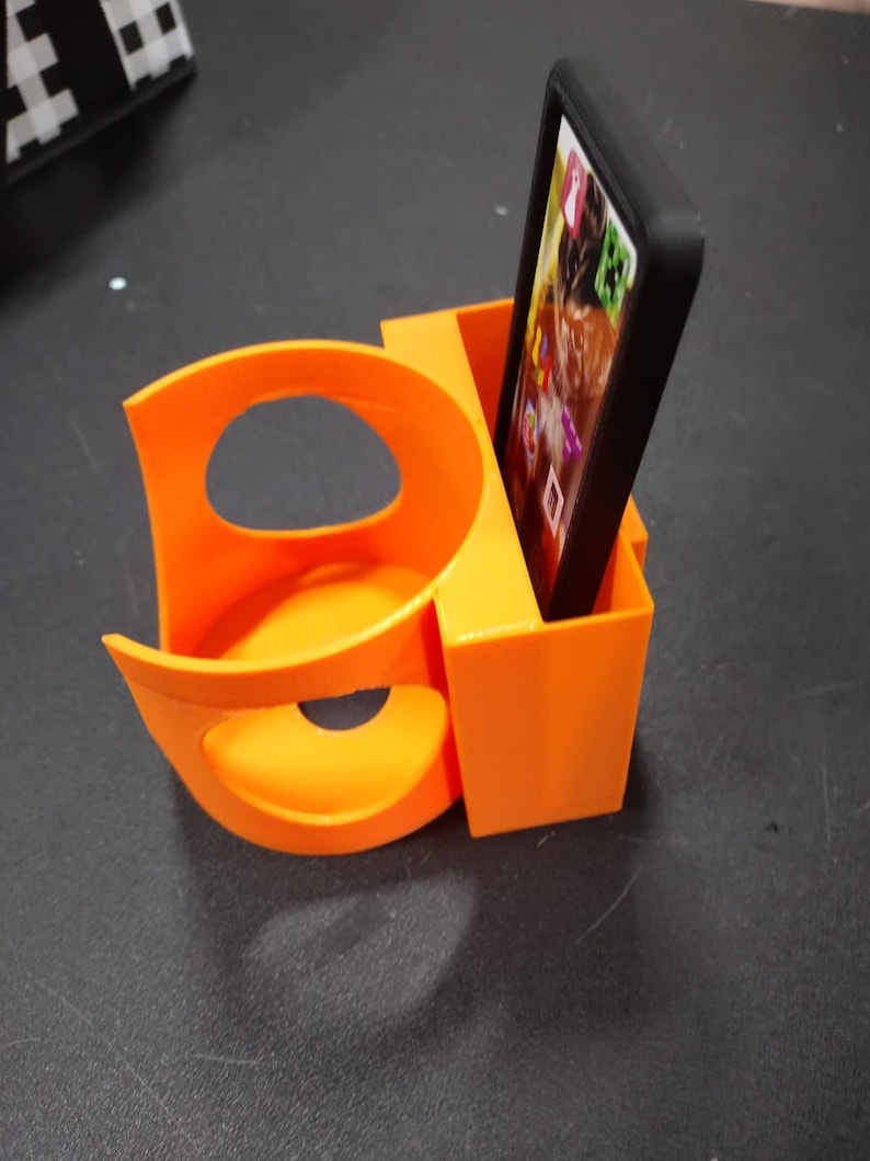 May include: Bright orange 3D-printed car cup holder and phone holder. The cup holder has a curved design with a circular opening. The phone holder is a rectangular box, holding a black phone with a screen displaying app icons.