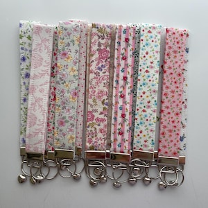 May include: A selection of fabric wrist keychains featuring floral designs in shades of pink, white, and blue. Each keychain has a silver-coloured metal clasp and a heart-shaped charm. The keychains are displayed in a row.