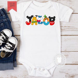 May include: White baby onesie with the name "JACOB" spelled out in colorful letters featuring Disney characters. The letters are outlined in black and have Mickey Mouse ears.