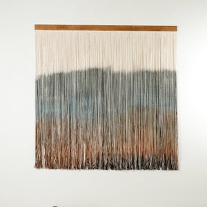 以下が含まれることがあります： 白から茶色へのグラデーションで糸で作られた壁掛け。糸が織り込まれて、質感のある外観になっています。壁掛けは木製の棒に掛けられています。