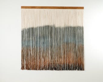 May include: A wall hanging made of yarn with a gradient of colors from white to brown. The yarn is woven together to create a textured look. The wall hanging is hung on a wooden dowel.