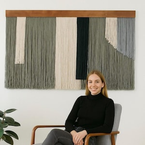 May include: A large, rectangular wall hanging with a wooden top bar. The textile art piece features vertical strands in shades of green, cream, black, and grey. The design includes geometric shapes and textures, creating a modern, abstract aesthetic.