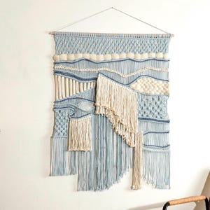 Peut inclure: Une grande tapisserie murale en macramé tissé avec un motif bleu et blanc. La tapisserie présente une variété de textures et de motifs, notamment des franges, des nœuds et des boucles.