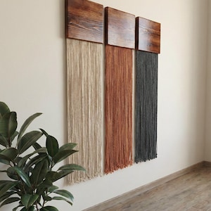 Op de afbeelding: Een moderne wanddecoratie met drie houten panelen met een getextureerde, donkerbruine afwerking. Elk paneel ondersteunt een andere kleur hangend garen: beige, roest en antracietgrijs. Het kunstwerk is op een neutrale muur gemonteerd.