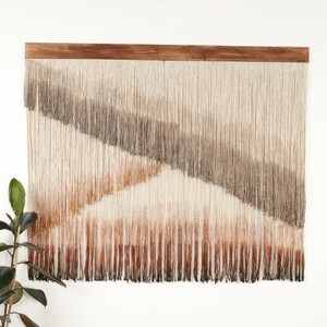 May include: A large, rectangular wall hanging with a wooden top. The woven fibers transition from light beige to brown, creating a diagonal pattern. The bottom edge has a fringe. A houseplant is visible on the left.