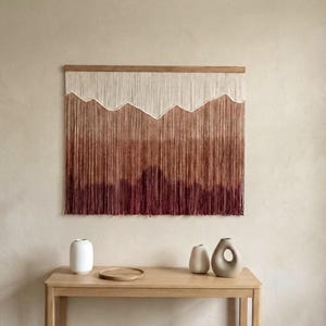 May include: A wall hanging with a mountain design, woven from threads in shades of off-white, brown, and burgundy. It hangs on a wooden frame. Beneath it, a wooden table holds white and beige vases, creating a minimalist aesthetic.