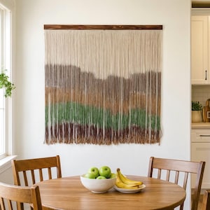 May include: A large, rectangular wall hanging with a wooden top bar. The hanging features vertical strands in shades of beige, brown, and green, creating a layered, textured effect. The piece is mounted on a white wall, above a wooden table.