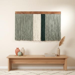 Peut inclure: Une tenture murale décorative avec un cadre en bois, composée de brins de fil verticaux verts, blancs et vert foncé. Un banc en bois en dessous supporte des vases et des livres. La pièce a une palette de couleurs neutres.