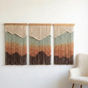 May include: Three wall hangings with a mountain design. Each piece has a wooden top and vertical strands in beige, green, orange, and brown, creating a gradient effect. The art pieces are displayed on a white wall, adding a touch of natural decor.