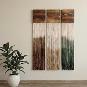 May include: Three rectangular wall hangings with wooden tops and fringed bottoms. The fringe transitions from white to brown, tan, and green. A potted plant with green leaves sits to the left of the wall art.