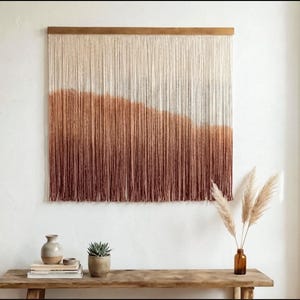 Può includere: Un grande arazzo da parete con una barra superiore in legno. L'arazzo presenta fili verticali di filato in un gradiente di colori, che vanno dal crema alle tonalità del marrone e del bordeaux. L'arazzo è esposto sopra una panca di legno.