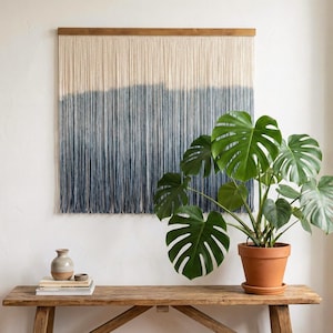 May include: A cream and blue ombre wall hanging with a wooden top. A Monstera plant in a terracotta pot sits on a wooden bench. The plant adds a natural element to the room's decor.