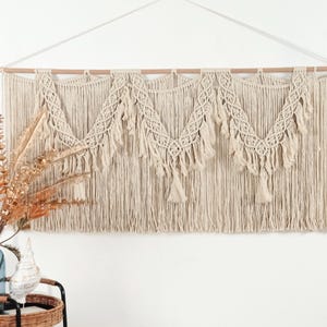 May include: A large, cream-colored macrame wall hanging. The piece features a wooden dowel at the top with intricate woven patterns and tassels. The design includes three decorative sections with a fringe of hanging cords, adding a bohemian touch to home decor.