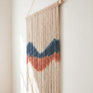 Può includere: Un arazzo da parete in macramè beige con un motivo a onde blu e arancioni. L'arredamento della parete è sospeso da un tassello di legno e pende contro un muro bianco. Il pezzo è un design moderno e minimalista.