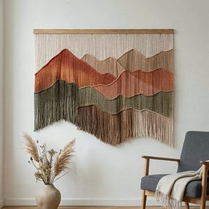 May include: A large, woven wall hanging depicting a mountain range in shades of orange, brown, and green. The artwork is mounted on a wooden bar and hangs against a white wall. The piece has a fringe at the bottom.