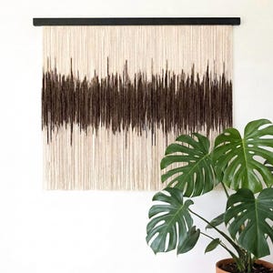 May include: A beige and brown fiber art wall hanging. The artwork features a dark brown, horizontal band of varying thickness against a beige background. The piece is suspended from a black wooden rod. A potted Monstera plant is in the foreground.