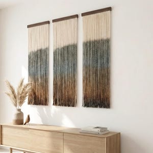 May include: Three wall hangings with a gradient color scheme, transitioning from beige to blue and brown. Each piece features a dark wooden top and long, fringed strands. The decor is displayed above a light wood cabinet with a vase and decorative bird.