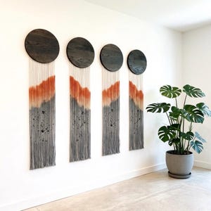 Könnte beinhalten: Vierteiliges Wandkunst-Set. Jedes Stück hat einen dunklen Holz-Kreis oben, mit einer langen, gefransten Tapisserie darunter. Die Wandbehänge gehen von Orange zu Grau über, mit schwarzen Akzenten. Rechts ist eine Topfpflanze zu sehen.