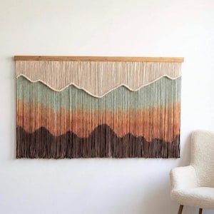May include: A large wall hanging with a mountain design. The top section is beige, transitioning to green, orange, and brown. The piece is made of yarn and mounted on a wooden bar. The overall design resembles a sunset over a mountain range.